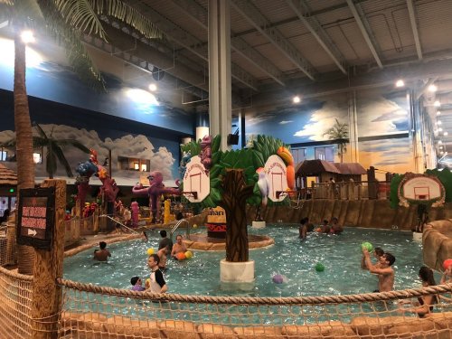 Indoor water park scene with children playing in a pool surrounded by tropical decorations and colorful water features.