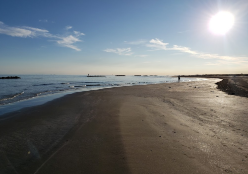A serene beach scene with gentle waves, a clear sky, and a person walking along the shore under a bright sun.
