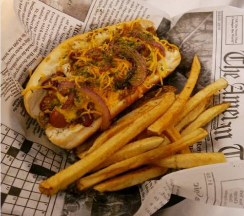 A loaded hot dog topped with cheese and onions, served with a side of golden French fries in a newspaper-lined basket.