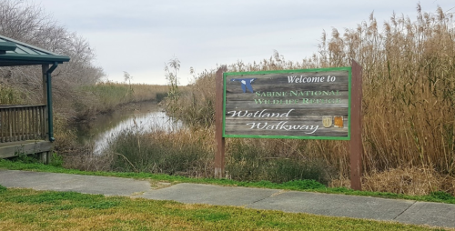 Sign welcoming visitors to Sabine National Wildlife Refuge, featuring a wetland walkway beside a calm waterway.