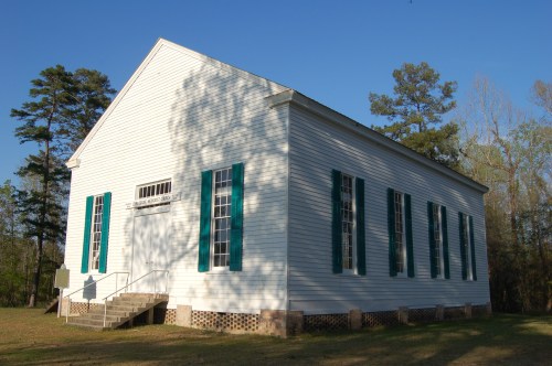 A white wooden church with green shutters, surrounded by trees and grass under a clear blue sky.