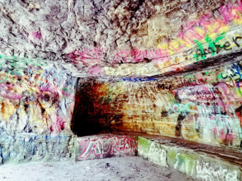 Colorful graffiti covers the walls of a rocky cave, with a curved stone bench in the foreground.