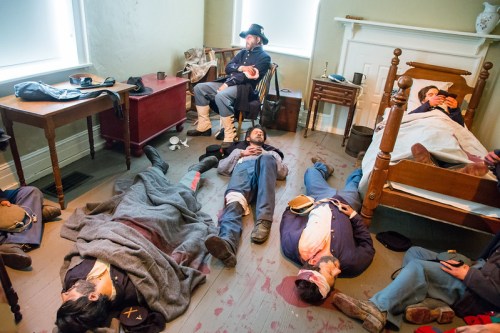 Civil War reenactors in a hospital setting, some lying on the floor and others resting on beds, depicting wartime injuries.