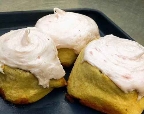 Three freshly baked cinnamon rolls topped with pink frosting, placed on a dark tray.
