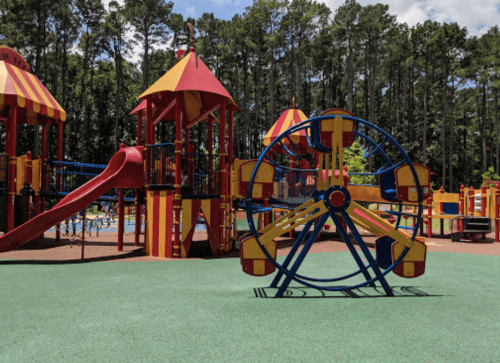 Colorful playground with a slide, climbing structures, and a small Ferris wheel, surrounded by trees and blue sky.