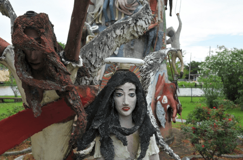 A surreal sculpture featuring figures with distorted features, surrounded by greenery and a cloudy sky.