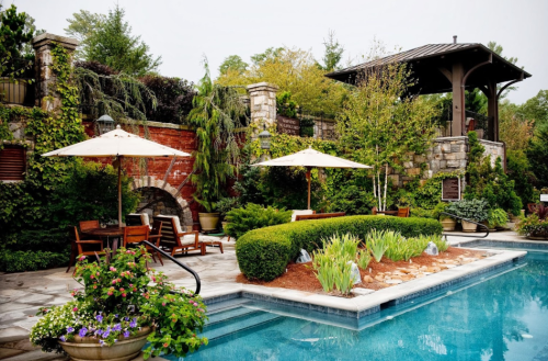 A serene pool area with lush greenery, lounge chairs, umbrellas, and a stone wall in a tranquil garden setting.