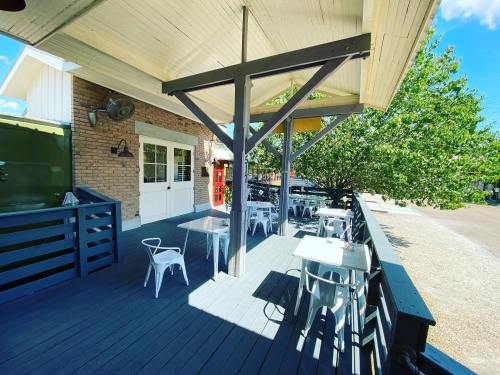 A sunny outdoor patio with white tables and chairs, surrounded by greenery and a brick building in the background.