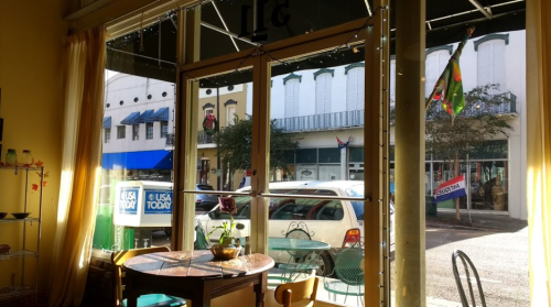 View from inside a café, showing a street with parked cars and storefronts, framed by large glass doors.