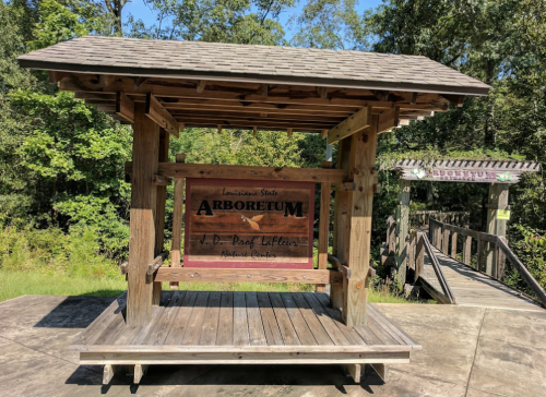 Wooden structure with a sign reading "Louisiana State Arboretum" surrounded by trees and a bridge in the background.