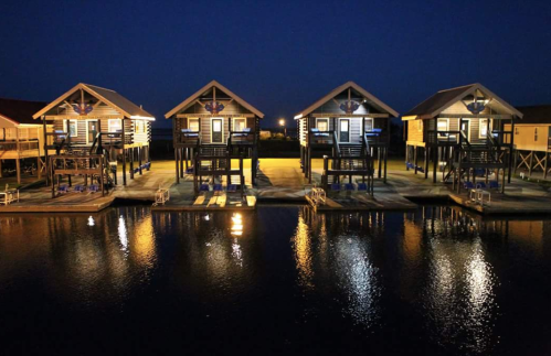 Four illuminated waterfront cabins at night, reflecting in calm water, with a serene atmosphere.