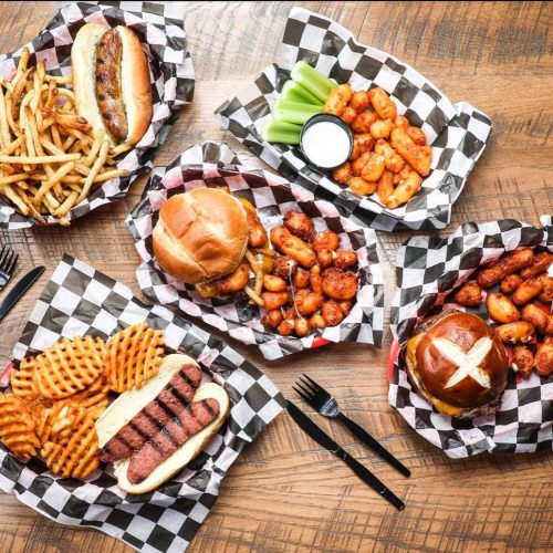 A spread of various foods including hot dogs, burgers, chicken wings, fries, and celery on checkered paper.
