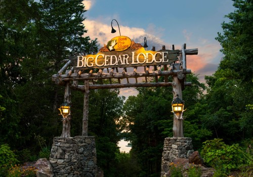 Sign for Big Cedar Lodge with wooden structure and lanterns, surrounded by trees under a colorful sky.