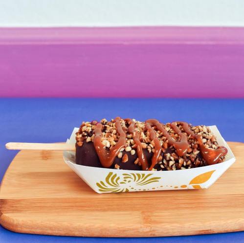 A chocolate-covered ice cream bar topped with nuts and drizzled with caramel, served in a decorative paper boat.