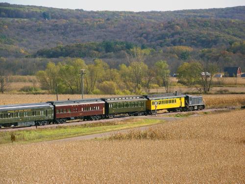 A vintage train with green, red, and yellow cars travels through a scenic landscape of fields and autumn trees.