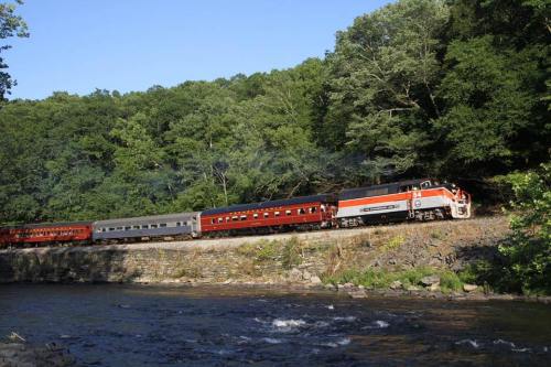 A vintage train travels along a riverbank, surrounded by lush green trees under a clear blue sky.