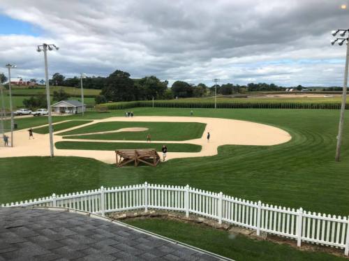 A scenic view of a baseball field with players, surrounded by green grass and a white picket fence under a cloudy sky.