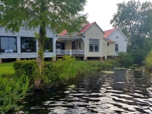 A waterfront house with a porch, surrounded by greenery and water, featuring a red roof and wooden siding.