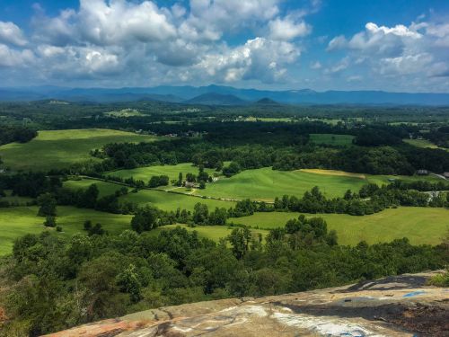 A panoramic view of lush green fields and rolling hills under a partly cloudy sky. Mountains are visible in the distance.