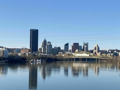 Pittsburgh skyline reflecting on the water, featuring tall buildings and a bridge under a clear blue sky.