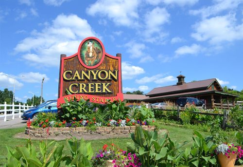 Sign for Canyon Creek Dining Steakhouse surrounded by colorful flowers and greenery under a blue sky.