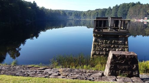 A calm lake reflects trees and a stone structure on the shore, surrounded by lush greenery and a clear blue sky.