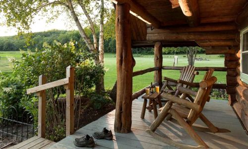 A cozy log cabin porch with two rocking chairs, a small table, and a view of a green field and trees.