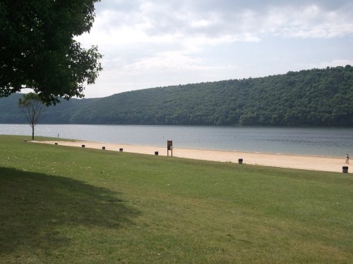 A serene lakeside view with green hills, a sandy beach, and a grassy area under a cloudy sky.