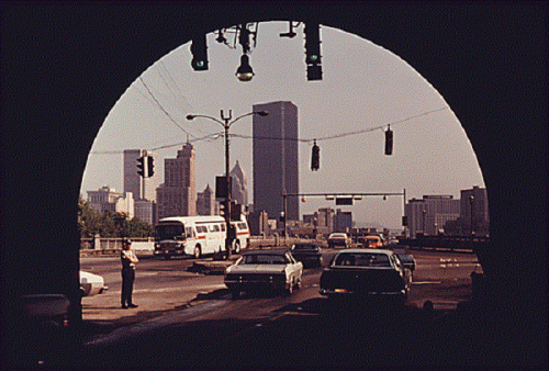 View of a city skyline through a tunnel, with cars and traffic lights visible in the foreground.