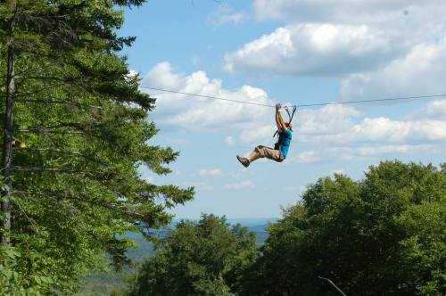 A person zip-lining through a forested area with a blue sky and clouds in the background.