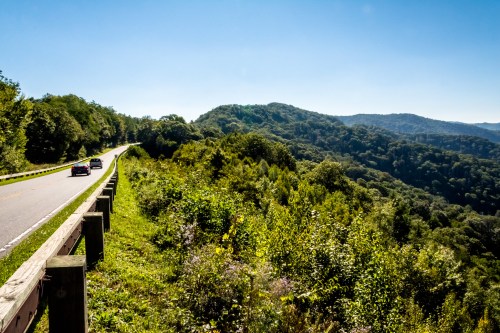 A scenic road winds through lush green hills under a clear blue sky. Cars drive along the roadside.