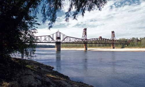 A rusted railway bridge spans a river, surrounded by trees and a clear blue sky with scattered clouds.