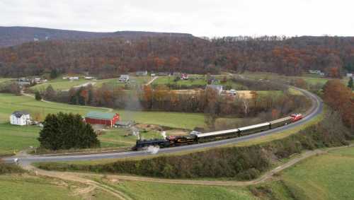 A steam train travels along a winding track through a rural landscape with autumn foliage and scattered houses.