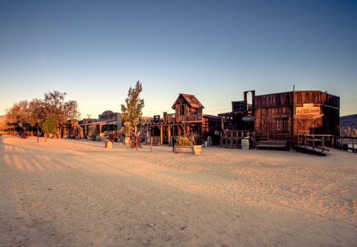 Desert landscape featuring rustic wooden buildings at sunset, with a sandy path and sparse vegetation.