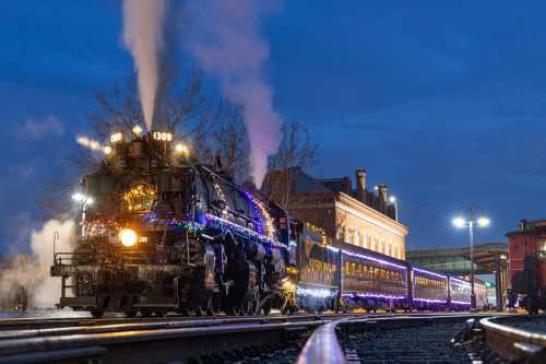 A vintage steam train adorned with colorful lights, billowing smoke, at dusk near a historic station.