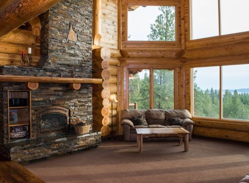Cozy wooden cabin interior with a stone fireplace, large windows, and a view of trees and mountains outside.