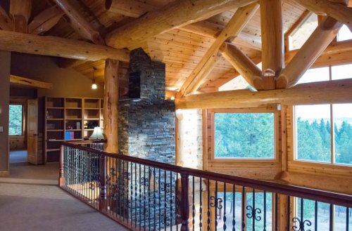 Interior of a log cabin with wooden beams, a stone fireplace, and large windows overlooking a forested landscape.