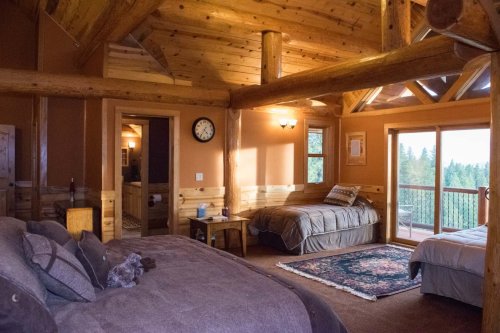 Cozy cabin interior with wooden beams, two beds, a rug, and a balcony overlooking a forested area.