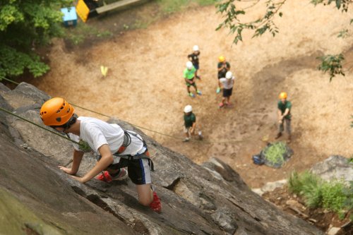 A climber ascends a rock face while others observe and prepare below in a forested area.