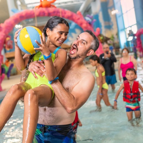 A man joyfully lifts a girl in a swimsuit while playing in a water park, surrounded by other children and adults.