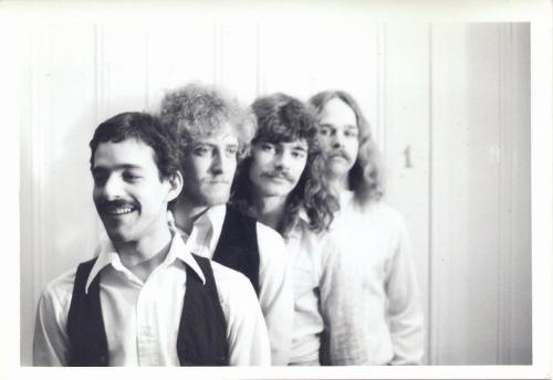 Four men with varying hairstyles and mustaches stand in a line against a white wall, smiling at the camera.
