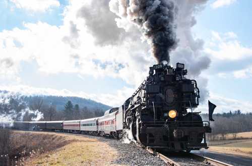 A vintage steam locomotive billows smoke as it travels along a scenic railway, surrounded by hills and a blue sky.