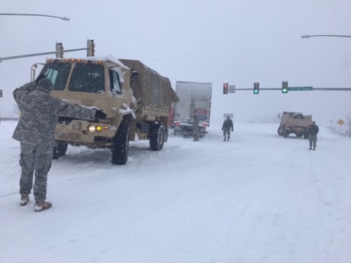 Military vehicles and personnel navigate a snowy road during a winter storm, with traffic signals visible in the background.