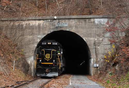 A black locomotive emerges from a stone tunnel surrounded by autumn foliage, with "BRUSH 1911" engraved above the entrance.