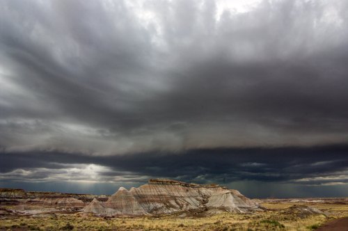 Dramatic dark clouds loom over a rugged, layered landscape, creating a striking contrast with the earthy tones below.