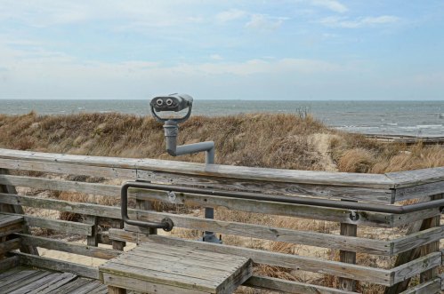 A viewing telescope on a wooden deck overlooking a sandy beach and the ocean under a cloudy sky.