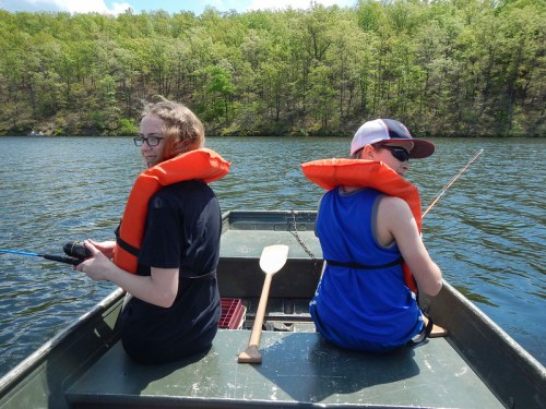 Two children in life jackets sit back-to-back in a small boat, fishing on a calm lake surrounded by green trees.