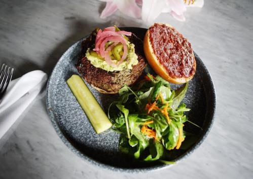 A gourmet burger with pickled onions and avocado on a plate, accompanied by a pickle and a side salad.