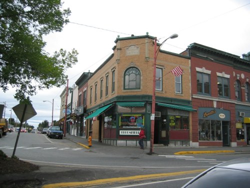 Historic buildings at a street corner, featuring storefronts and an American flag, under a cloudy sky.