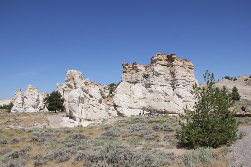 White rock formations rise against a clear blue sky, surrounded by sparse vegetation and a few visitors walking nearby.